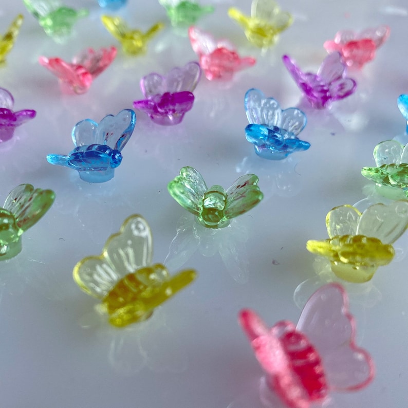May include: A collection of small, translucent butterfly-shaped embellishments in various colors, including pink, blue, green, yellow, and purple. The butterflies are scattered across a reflective surface, creating a colorful and whimsical display.