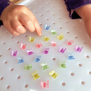 May include: A collection of small, colorful butterfly-shaped beads in shades of pink, purple, yellow, green, and blue. The beads are scattered on a white surface, with a child's hand reaching towards them.