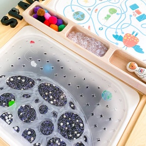Könnte beinhalten: Ein hölzerner Aktivitätstisch mit einem durchsichtigen Plastiktablett gefüllt mit violettem und weißem Sinnesreis. Das Tablett hat einen mondförmigen Ausschnitt mit kleineren Kreisen darin. Es gibt ein separates Fach mit bunten Pompons und ein Fach mit durchsichtigen Plastikperlen. Der Tisch hat ein Schild mit der Aufschrift "SPACE" in schwarzen Buchstaben.
