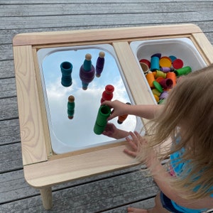 May include: A wooden sensory table with two white plastic bins. One bin has a mirrored surface, reflecting colorful wooden toys. The other bin is filled with various wooden toys. A child is playing with the toys.