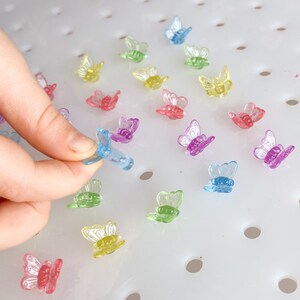 May include: A collection of colorful, translucent butterfly-shaped craft embellishments. The butterflies are in shades of pink, purple, yellow, green, and blue. One butterfly is being held by a hand. The butterflies are on a white surface.