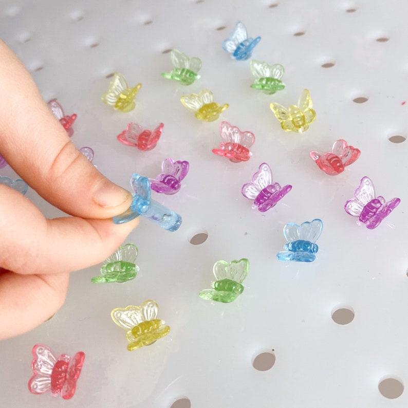 May include: A collection of colorful, translucent butterfly-shaped craft embellishments. The butterflies are in shades of pink, purple, yellow, green, and blue. One butterfly is being held by a hand. The butterflies are on a white surface.