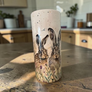 May include: A cylindrical tumbler featuring a cream-coloured background with black speckles. The surface displays a detailed illustration of two brown hares standing amidst a field of wildflowers and tall grasses, with a small blue bird in flight.