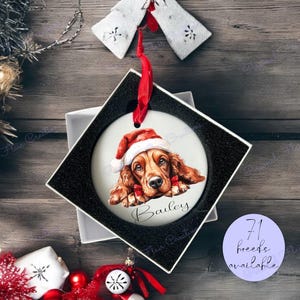 May include: A white ceramic Christmas bauble with a red ribbon, featuring a watercolour illustration of a dog wearing a Santa hat. The dog is a red and white Irish Setter with a bow tie. The bauble is personalised with the name "Bailey".