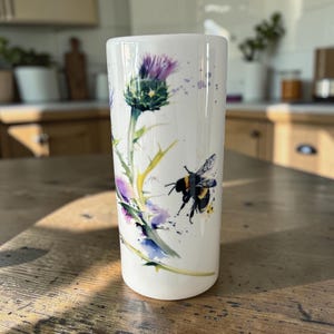 May include: A white cylindrical ceramic tumbler featuring a watercolour illustration of a purple thistle flower and a black and yellow bumblebee. The design includes green stems and leaves with subtle purple and blue paint splatters on the surface.