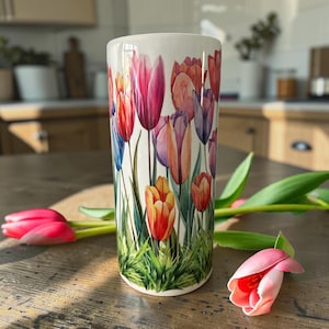 May include: A cylindrical ceramic tumbler featuring a vibrant watercolour print of multi-coloured tulips in shades of red, orange, pink, and purple, set against a white background with green stems and leaves at the base of the design.