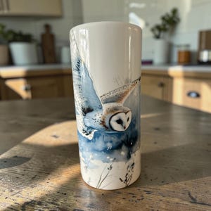 May include: A cylindrical white ceramic vase featuring a detailed illustration of a barn owl in flight. The owl has white and brown feathers, and the background depicts a soft blue and white watercolour-style winter landscape with dark silhouettes.