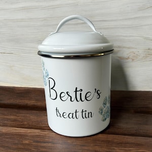 May include: A white cylindrical metal treat tin with a matching lid and a top handle. The front features black cursive text that reads Bertie's treat tin, accented by two blue floral-patterned paw prints on the left and right sides of the text.