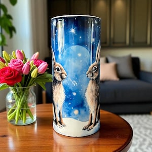 May include: A tall, cylindrical vase depicting two hares against a blue night sky with a full moon and stars. The vase is placed on a wooden table, alongside a vase of flowers. The vase's height is approximately 25 cm.