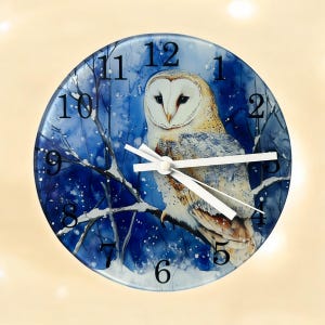 May include: A round glass clock featuring a white barn owl perched on a snowy branch. The clock face has black Roman numerals and white hands. The background is a blue watercolour wash with white snowflakes.
