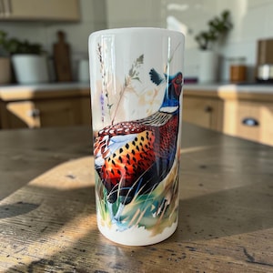 May include: A cylindrical white ceramic tumbler featuring a detailed illustration of a male pheasant with vibrant red, blue, and brown plumage. The bird is depicted standing among tall grasses and wildflowers, rendered in a painterly style.