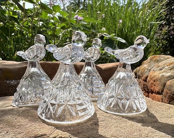 Puede incluir: Una colección de cinco figuritas de pájaros de cristal transparente. Cada pájaro se asienta sobre una base facetada en forma de cono. Los pájaros se exhiben sobre una gran superficie rocosa texturizada, con vegetación en el fondo.