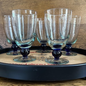 May include: Set of six clear glass goblets with a blue base and stem, arranged on a wooden tray with a black metal rim. The goblets have a classic design, perfect for serving beverages.