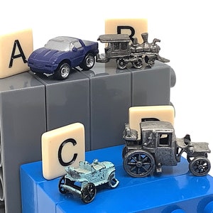 May include: A set of four miniature toy cars, including a blue sports car, a silver train engine, a silver vintage car, and a blue vintage car. The cars are arranged on a gray and blue Lego base with letter tiles spelling out "A, R, C".