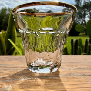 May include: A clear glass tumbler with a gold rim. The glass has a diamond-cut pattern on the lower half. The tumbler is sitting on a wooden surface, with a blurred green background.