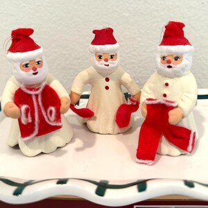 May include: Three ceramic Santa Claus figurines. Each Santa wears a red hat and holds a red garment. The figurines have white beards and are dressed in cream-colored robes. The figurines are on a white and green decorative surface.