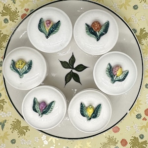 May include: A set of seven white ceramic containers with floral designs. Each container has a different floral design with green leaves and yellow, pink, and orange flowers. The containers are arranged in a circle on a white plate with a green leaf design in the center.