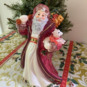 May include: A ceramic Santa Claus figurine, dressed in a burgundy coat with gold trim and a white robe. The figurine carries a sack and a basket with a lamb and a teddy bear. The Santa has a white beard and is decorated with festive details.