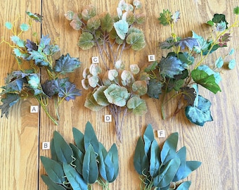 May include: Six artificial plant stems labeled A through F. Stems A, C, D, and E feature clusters of rounded, variegated green and purple leaves. Stems B and F consist of smooth, solid green, pointed leaves arranged in dense, upright groupings.