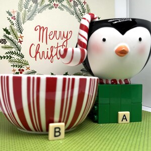 May include: A ceramic penguin-shaped mug with a candy cane handle and a red and white striped bowl. The mug sits on a green block base, and the bowl has vertical red and white stripes. The background features a Christmas wreath and the words "Merry Christmas".