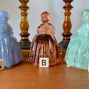 May include: Three glass figurines of women in different colors: light blue, pink, and green. The figurines are standing on a wooden surface with letter tiles in front of them, labeled A, B, and C.