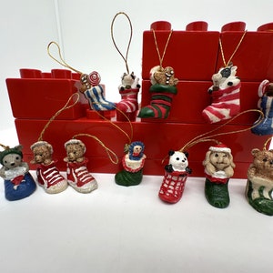 May include: A collection of 12 Christmas stocking ornaments featuring various animal and holiday themes. The ornaments are made of ceramic and have a painted finish. The ornaments are displayed on a red brick background.