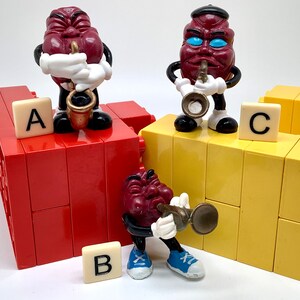 May include: Three red grape figurines playing musical instruments. The figurines are standing on top of yellow and red building blocks. The blocks are labeled with the letters A, B, and C.