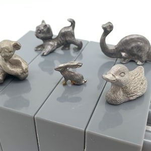 May include: A set of six miniature pewter animal figurines. The figurines include a koala, a cat, a rabbit, a duck, and a dinosaur. The figurines are displayed on a gray block surface.