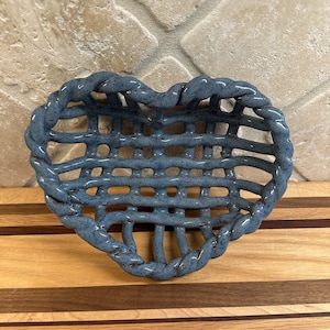 May include: A blue ceramic heart-shaped basket with a woven design. The basket is made of ceramic and has a textured, speckled appearance. The basket is sitting on a wooden cutting board.