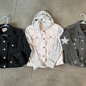 May include: Three denim jackets on hangers. A black denim jacket, a light pink jacket with a gray hooded sweatshirt attached, and a gray denim jacket with star and badge designs.