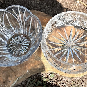 May include: Two clear glass bowls with intricate cut designs. The bowl on the left has a scalloped edge and a starburst pattern in the center. The bowl on the right has a circular shape with a starburst pattern.