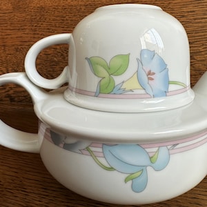 May include: A white porcelain teapot with a floral design featuring blue, yellow, and green flowers. The teapot has a lid and a handle. The teapot is sitting on a wooden surface.