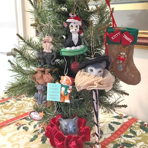 May include: A decorated Christmas tree with various cat-themed ornaments. Ornaments include a black and white cat wearing a Santa hat, a cat in a ruffled collar, and a stocking that says "Kitty Treats". Some ornaments have the text "A Friendly Christmas Greeting 1992".