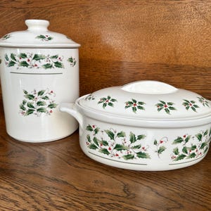 May include: Two white ceramic serving dishes with lids. The dishes are decorated with green holly leaves and red berries. One is a tall canister, the other is an oval casserole dish.