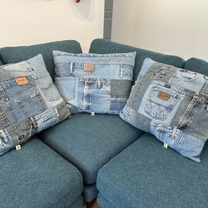 May include: Three square throw pillows made from repurposed denim jeans. Each pillow features a patchwork design with various shades of blue denim, including pockets, seams, and labels. The pillows are arranged on a teal blue sofa.