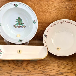 May include: Three white ceramic bowls with gold trim. The bowl on the left has a red poinsettia design. The bowl in the center has a gold trim and is blank. The bowl on the right has a red and green holly design and the text "Live Well, Laugh Often, Love Much".