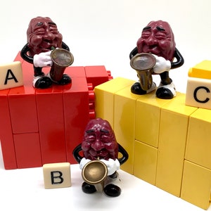 May include: Three purple grape figurines playing musical instruments. Each figurine is holding a different instrument: a trumpet, a saxophone, and a trombone. The figurines are standing on a yellow and red block structure. The blocks are labeled with the letters A, B, and C.