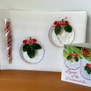 May include: Two white dreamcatchers with green leaves and red ribbons with the words "Hope" and "Peace" on them. A white card with the words "Legend of the Dreamcatcher" and a picture of two dreamcatchers with the words "Hope" and "Peace" on them. A red and white striped pen.