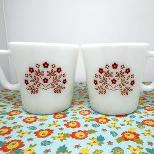 May include: Two white ceramic mugs with a red floral design. Each mug has a curved handle and a flared shape. The floral design features small flowers and leaves. The mugs are on a floral patterned tablecloth.
