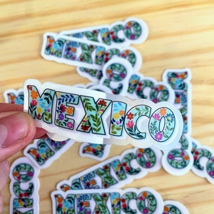 May include: A collection of stickers with the word "MEXICO" in a floral design. The letters are light blue with colorful flowers and green leaves. The stickers are on a light wood surface.