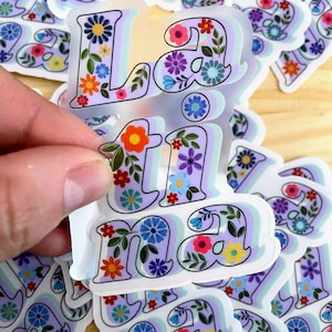 May include: A stack of colorful stickers with the word "Latina" in a floral design. The stickers are on a light brown surface.