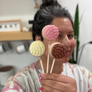 May include: A person holding four concha-shaped cake pops on sticks. The cake pops have different colors and patterns, including pink, brown, and yellow. The person is wearing a tie-dye shirt.