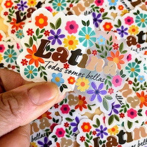 May include: A collection of floral stickers with the word "Latina" in brown letters, surrounded by colorful flowers. The text "Todas somos bellas" is also present. The stickers have a white scalloped edge and are scattered on a wooden surface.