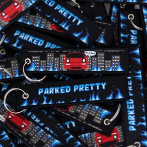 May include: A black keychain with blue flames and the text "Parked Pretty" in blue. The keychain features a red car in front of a city skyline with the text "This is fine" in a speech bubble above the car.