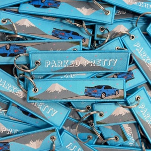 May include: A stack of blue and gray keychains with a white mountain and a blue car on them. The keychains have the text "Parked Pretty" on them.