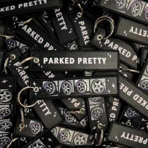 May include: A black and purple keychain with the text "Parked Pretty" and a graphic of four car wheels.