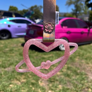 May include: A pink glittery heart-shaped handle with a Saturn-like ring and a small central heart, attached to a metallic rose gold strap featuring a pink cherry blossom flower emblem and a white tag with the text Parked Pretty.