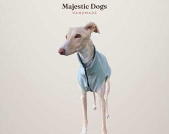 May include: A light-colored dog wearing a pale blue fleece vest with a front zipper. The text Majestic Dogs HANDMADE is printed in dark brown at the top. The dog has a slender build, pointed ears, and a light-colored coat.