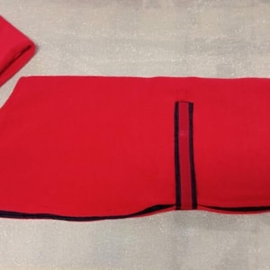 Könnte beinhalten: Roter Fleece-Hundemantel mit dunkelblauem Besatz und einem kleinen dunkelblauen Patch an der Seite. Der Mantel ist in der Hälfte gefaltet und ein passender roter Fleece-Halswärmer ist im Lieferumfang enthalten.