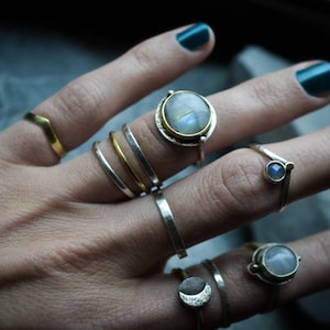 Puede incluir: Una mano con una variedad de anillos de plata y oro, incluyendo un anillo con una piedra de luna grande, un anillo con una piedra de luna más pequeña y un anillo con una pequeña piedra azul.